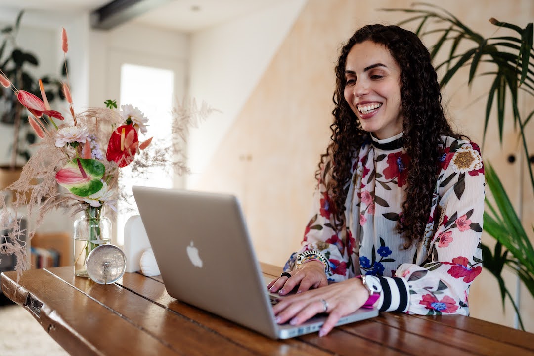 Monica of Dandelion Celebrations sits behind her laptop, smiling. She is wearing a floral print shirt.